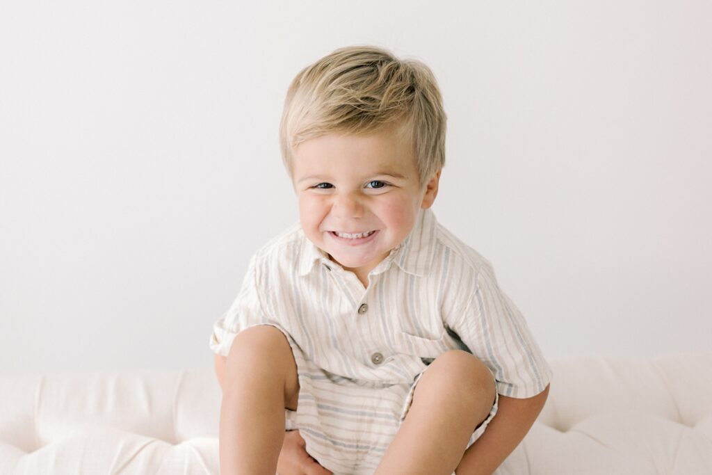 A little boys giggles while sitting on a covered linen bench in a white studio.