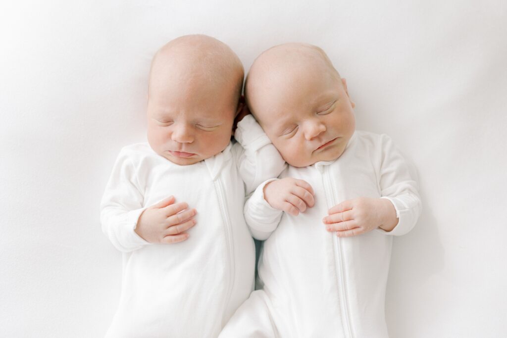 Twin boys sleep peacefully next to each other wearing matching white sleepers.