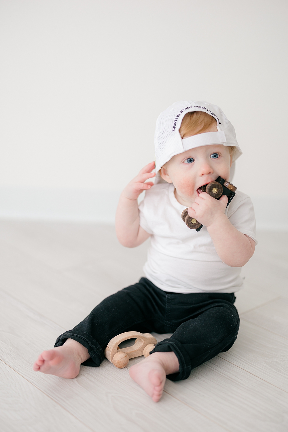 A little boy chews on a wooden toy car while he sits wearing a backwards white baseball cap.
