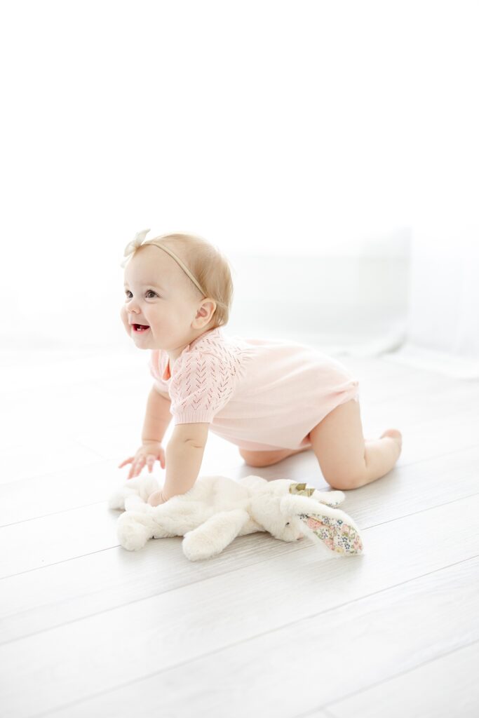 A little girl in a pink knit romper crawls across the floor with her white stuff bunny.