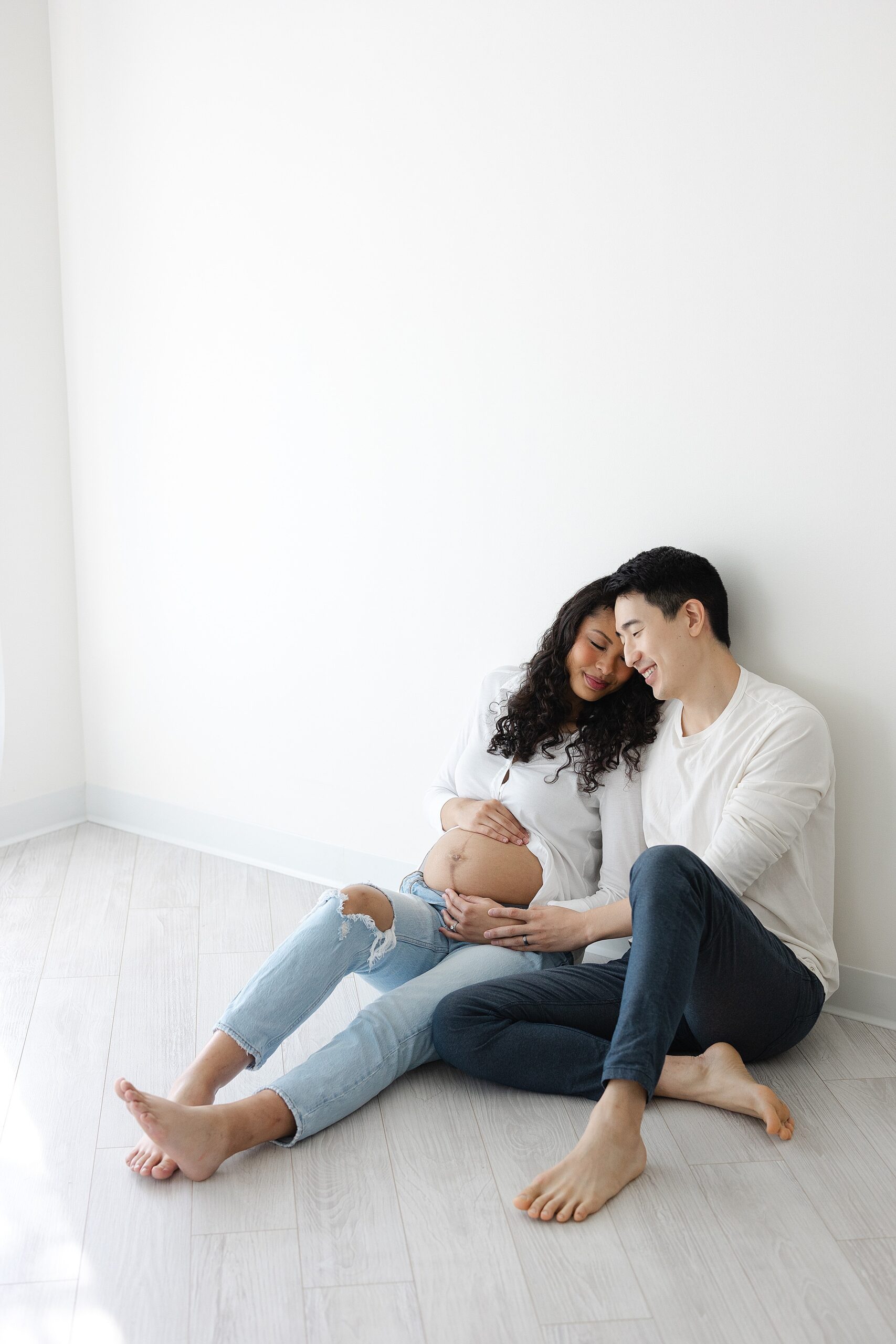 Happy expecting parents hold the bump while snuggling against a wall on the floor in jeans and white shirts after visiting Indianapolis Birthing Centers