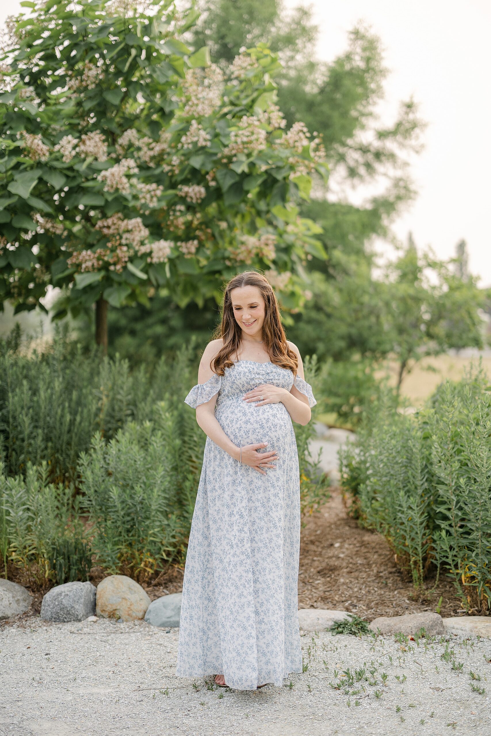 A pregnant woman in a floral print dress holds her bump while standing in a garden path