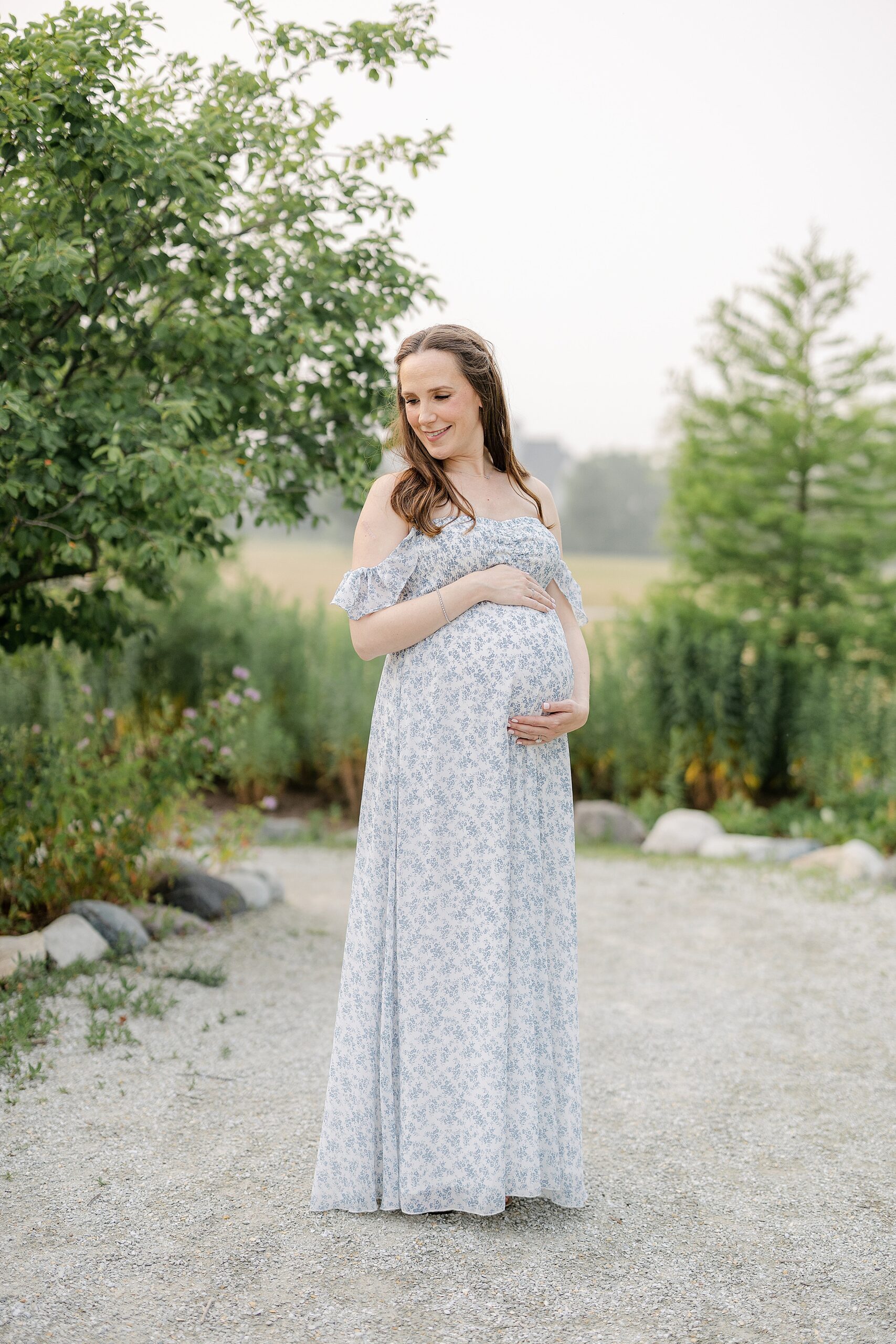 A mother to be in a blue floral print dress smiles down her shoulder while standing in a garden path at sunset before visiting Indianapolis Doulas