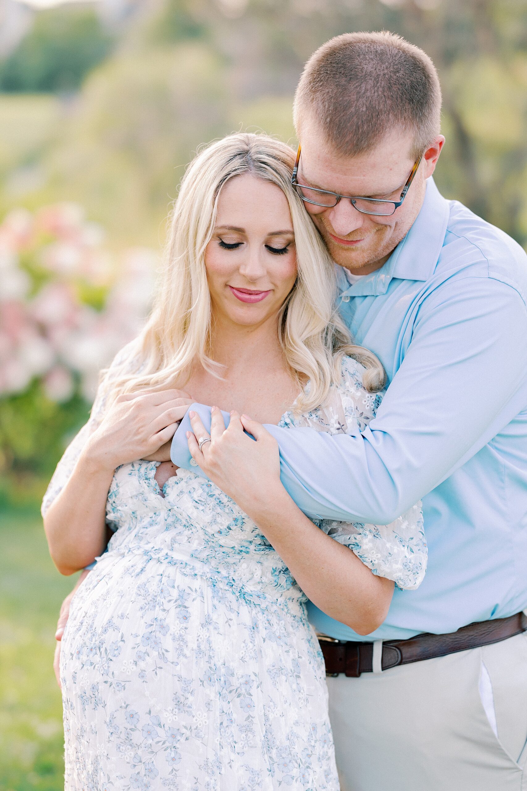 A mother to be leans into the chest of her husband in a blue floral print dress in a park at sunset