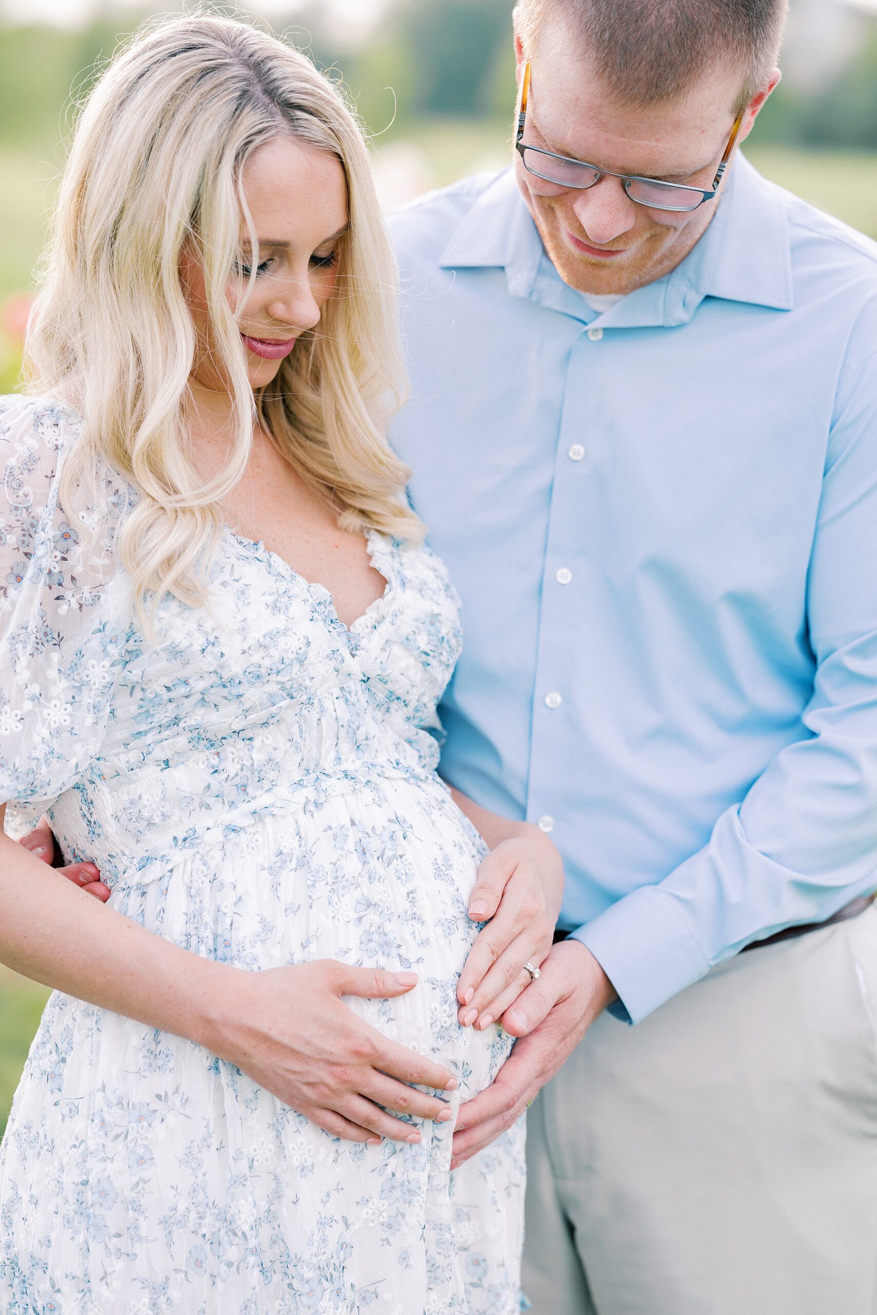 A father to be in a blue shirt smiles down to his hand on the bump with mom's hands in a floral print blue dress before meeting Indianapolis Midwives