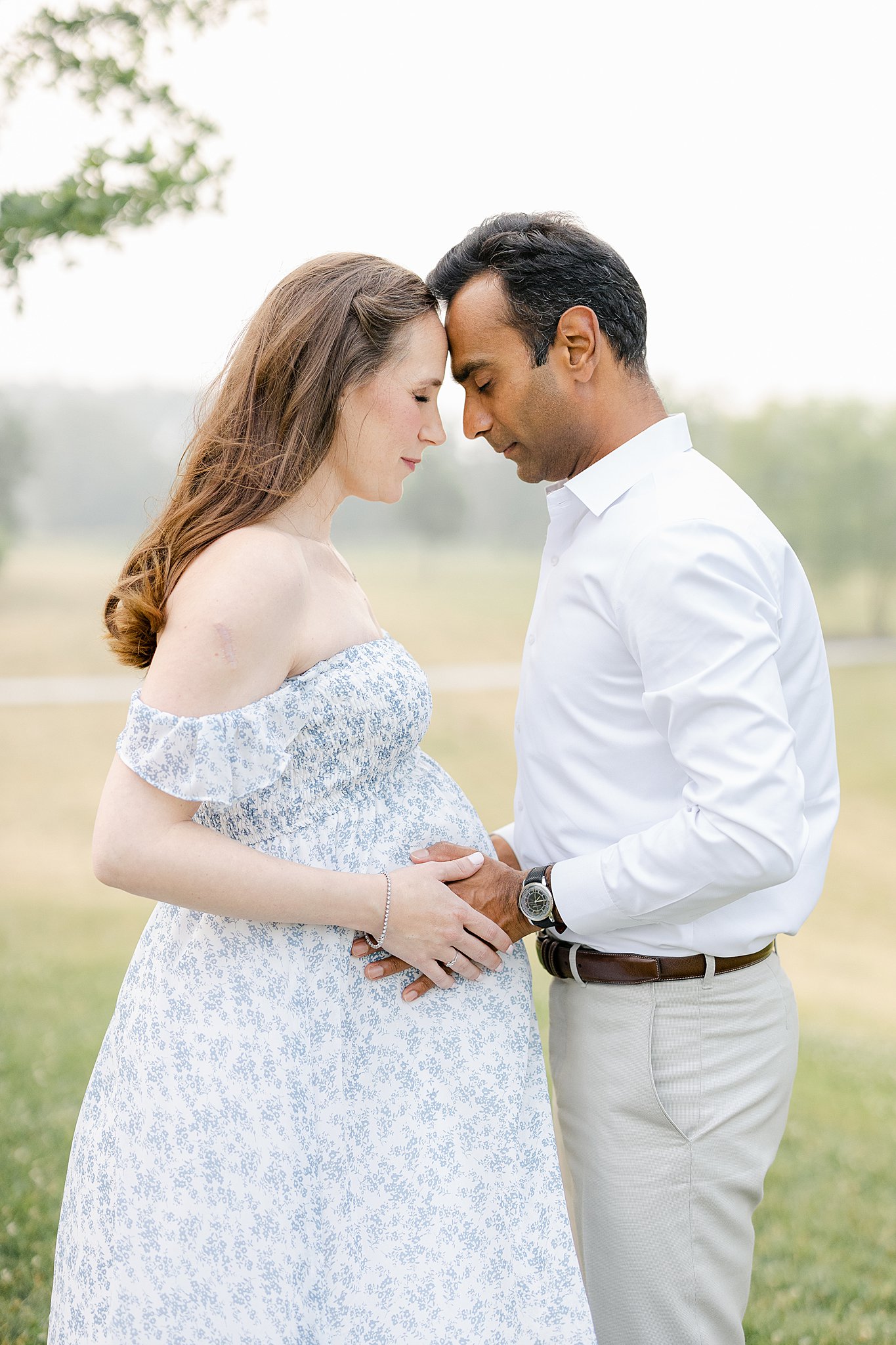 A pregnant mother and father touch foreheads while holding onto the bump and standing in a park field at sunset before a 3D Ultrasound Indianapolis