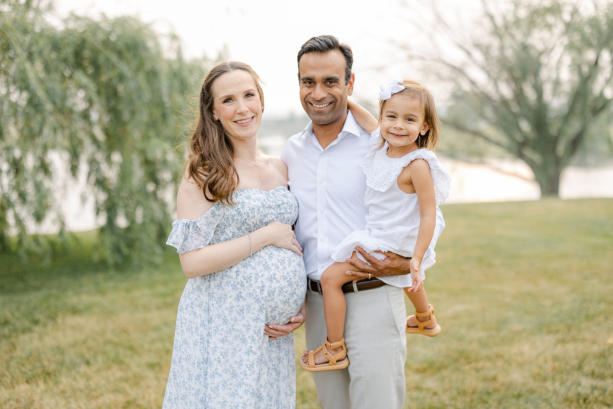 Happy expecting parents stand in a grassy field with their toddler daughter on dad's hip before getting a 3D Ultrasound Indianapolis
