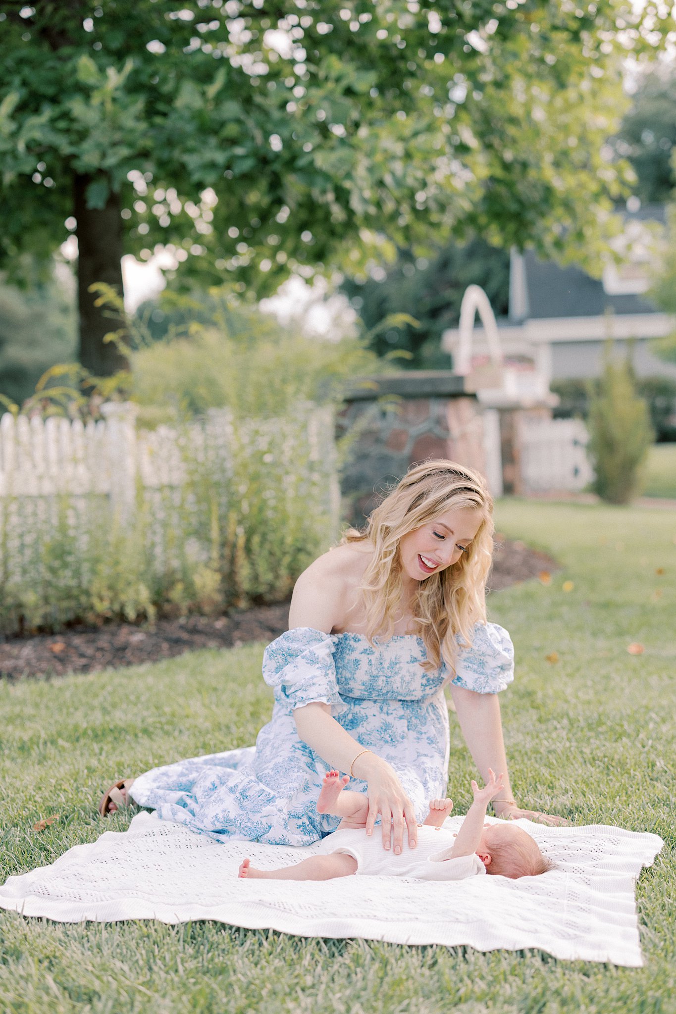 A happy new mom tickles her newborn baby laying on a picnic blanket in a garden