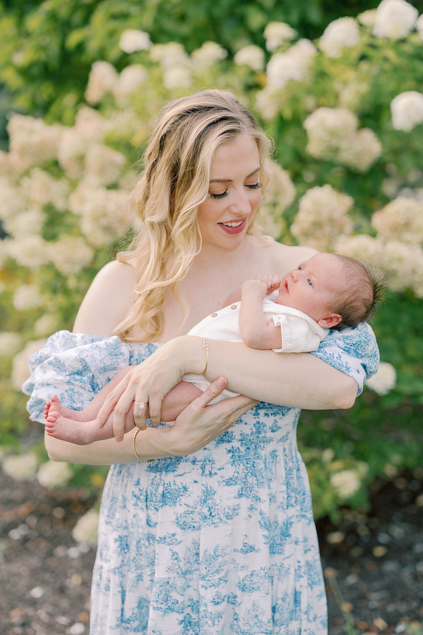 A happy mother in a blue dress stands in a garden holding her newborn baby in her arms in a white onesie thanks to a Fertility Clinic Indianapolis