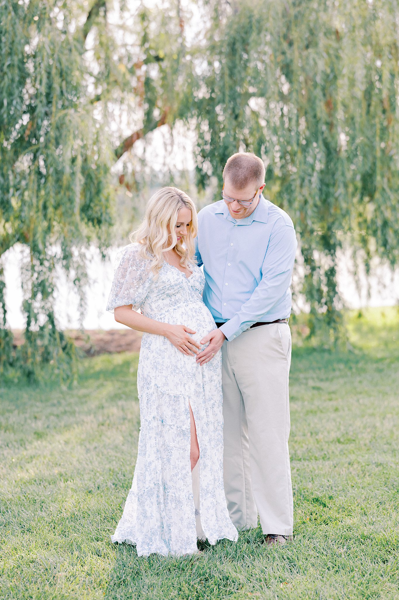 A mom to be in a floral print dress smiles down at the bump in her hands while standing lakeside under a willow tree