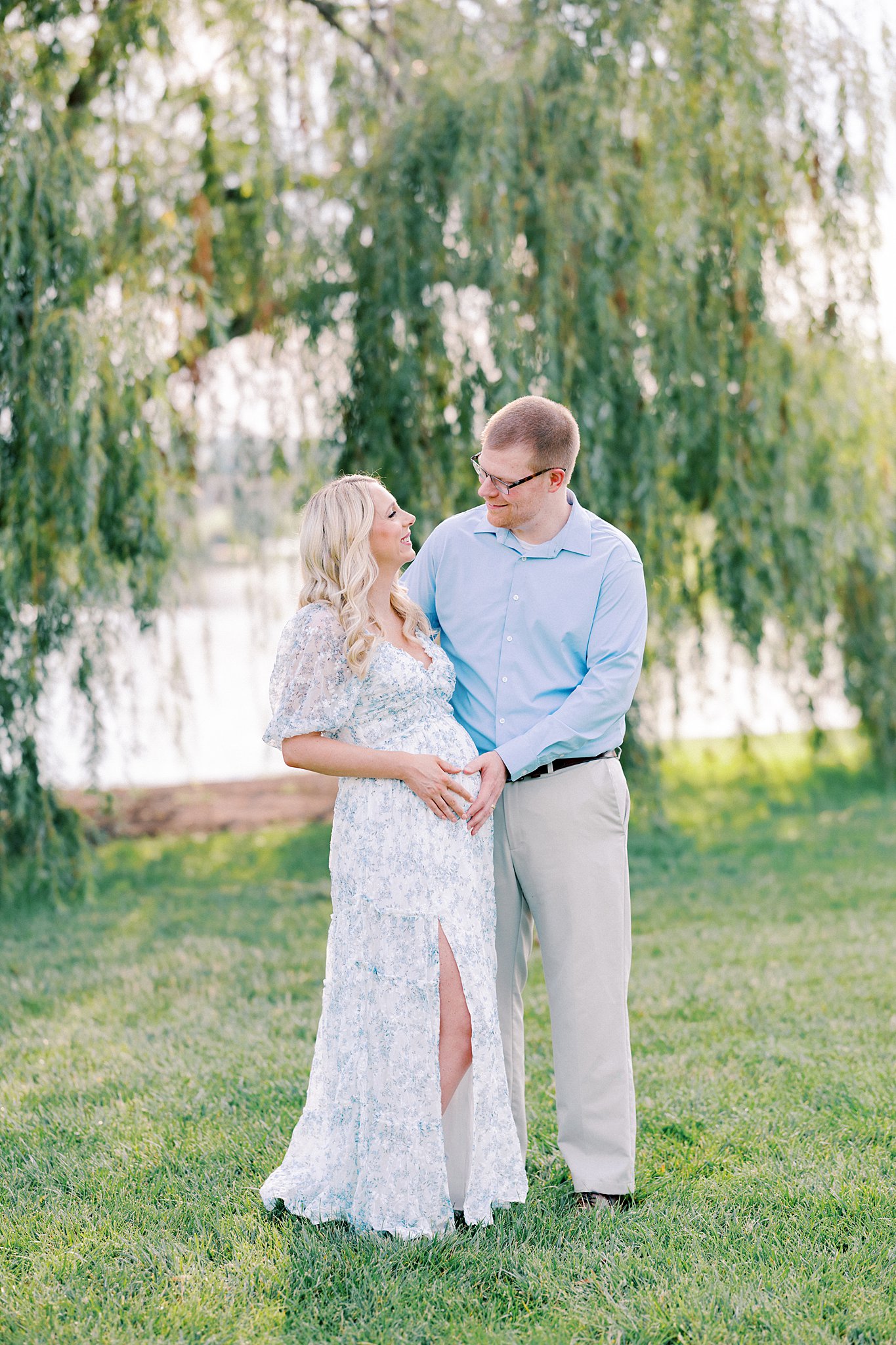 A mom and dad to be stand under a willow tree on a lake smiling at each other and holding the bump