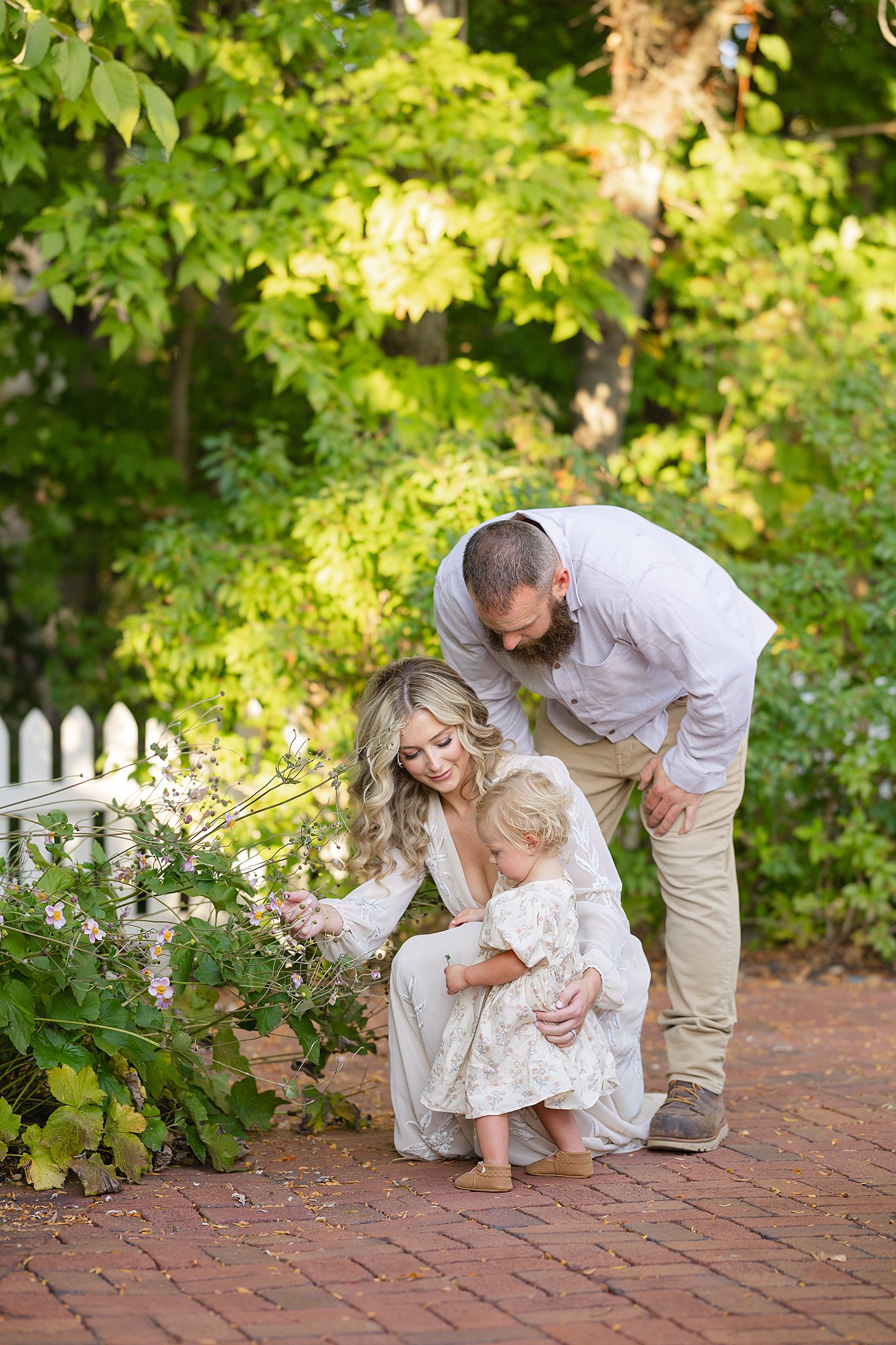 A mom and dad explore a garden and some pink flowers with their young toddler daughter