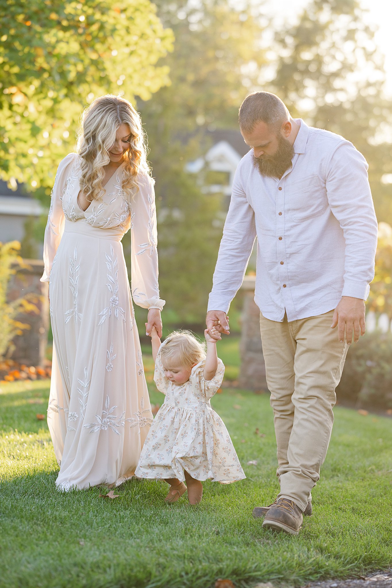 Happy parents walk through a garden lawn at sunset holding hands with their toddler daughter in a dress thanks to Indianapolis OBGYN