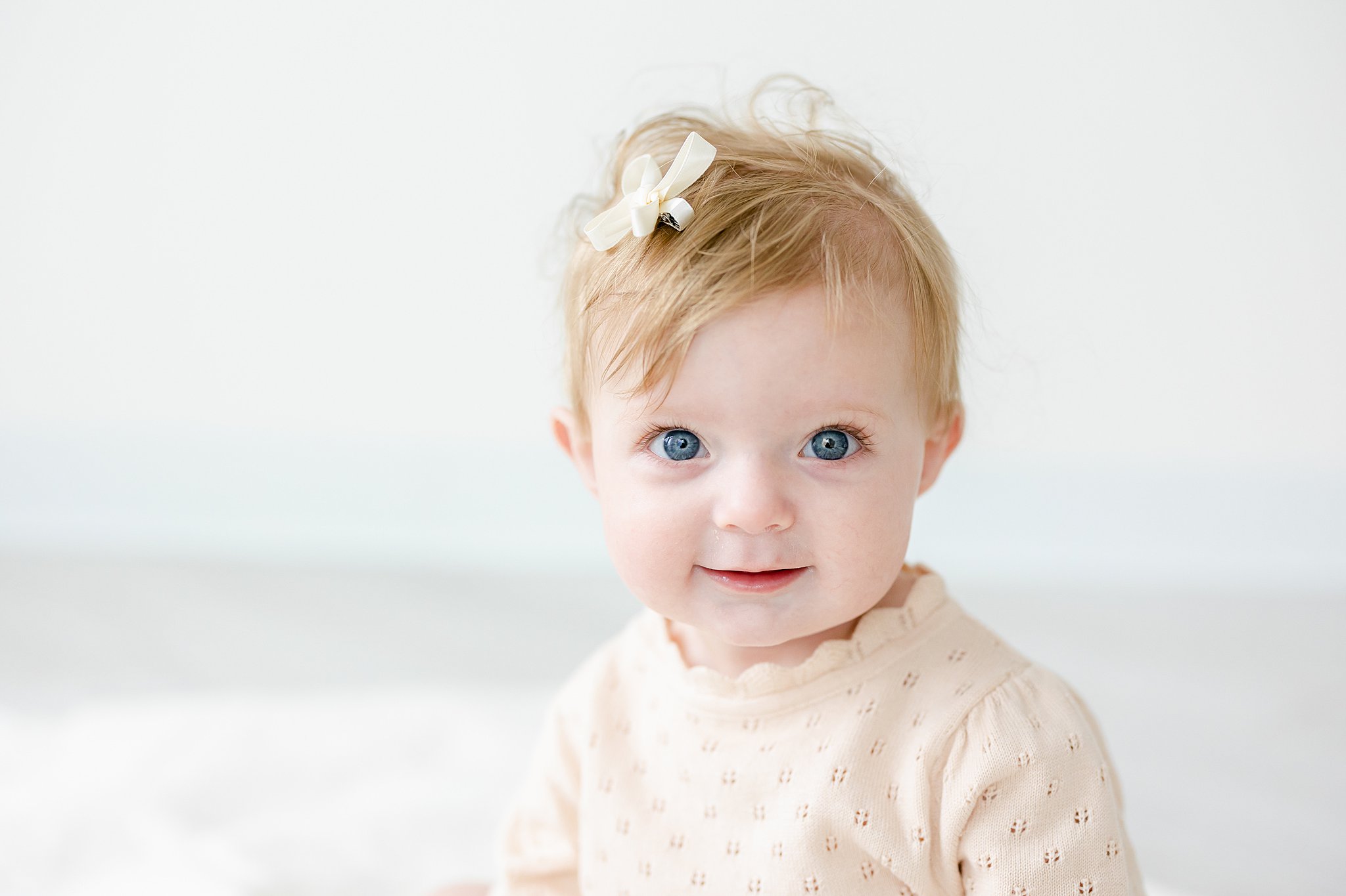 A toddler girl in a yellow onesie and hair bow sits in a studio smiling with big blue eyes