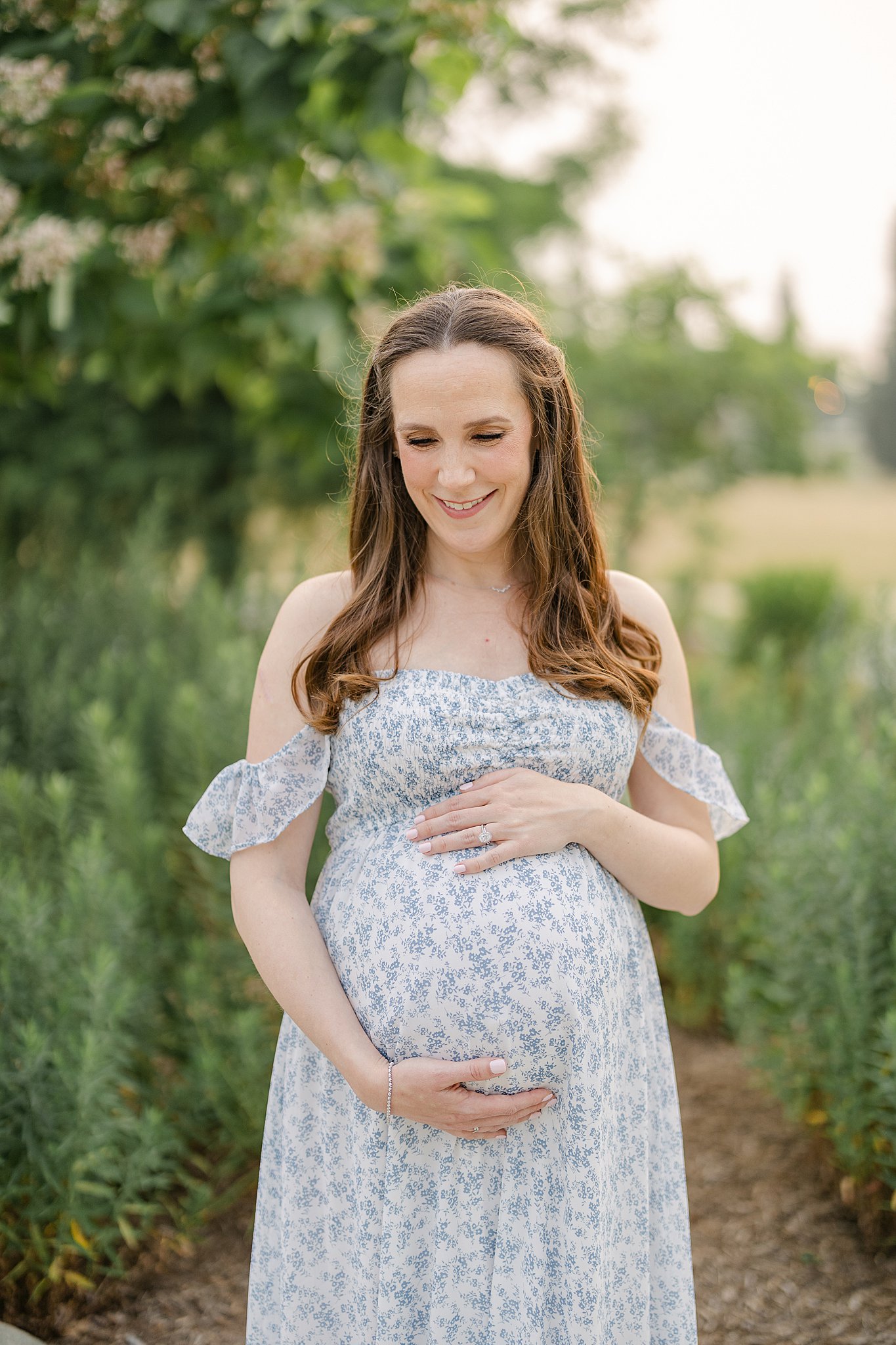 A happy expecting mom stands in a park trail holding her bump in a blue floral print dress after some Indianapolis prenatal yoga