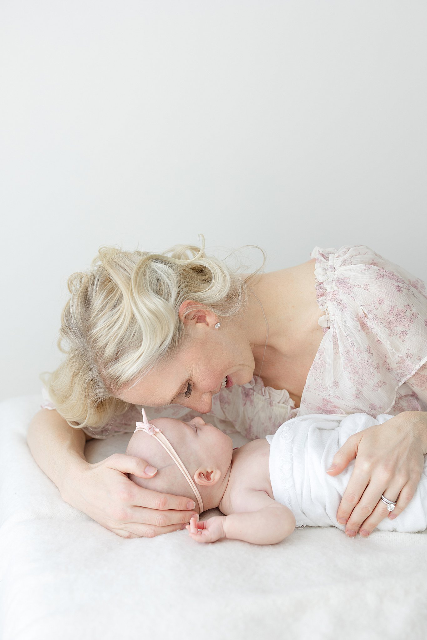 A happy mom laughs while snuggling with her sleeping newborn baby on a white bed in a pink dress after meeting lactation consultants in indianapolis