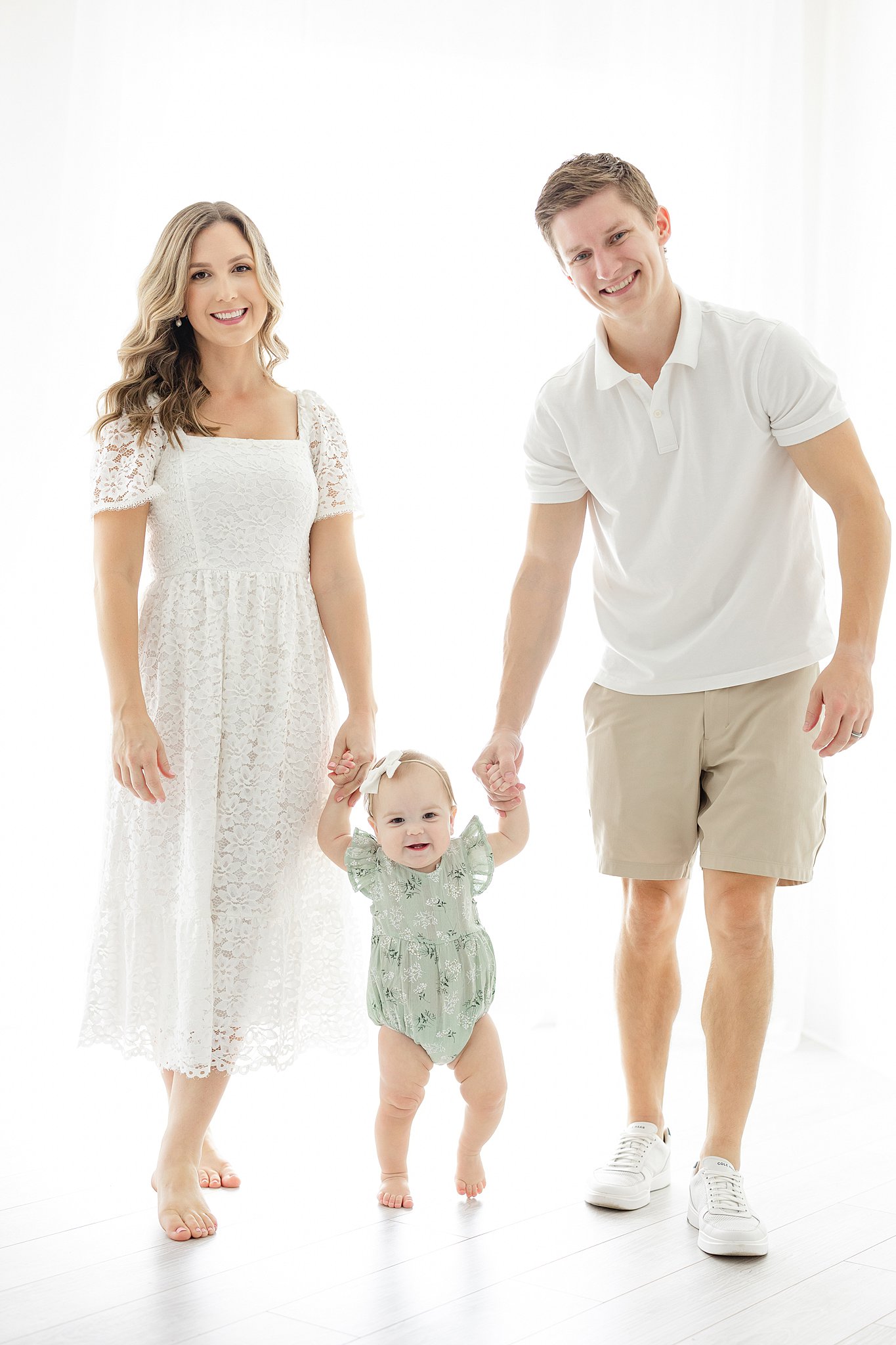 A mom and dad wearing white walk their toddler daughter through a studio holding her hands in a green onesie after meeting Nannies Indianapolis