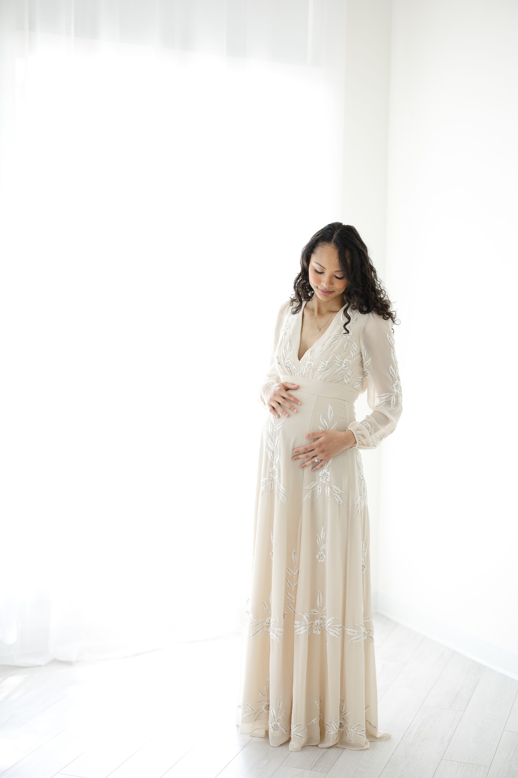 A mother to be in a cream maternity gown smiles down to her bump while standing in a studio before visiting a Prenatal Chiropractors in Indianapolis