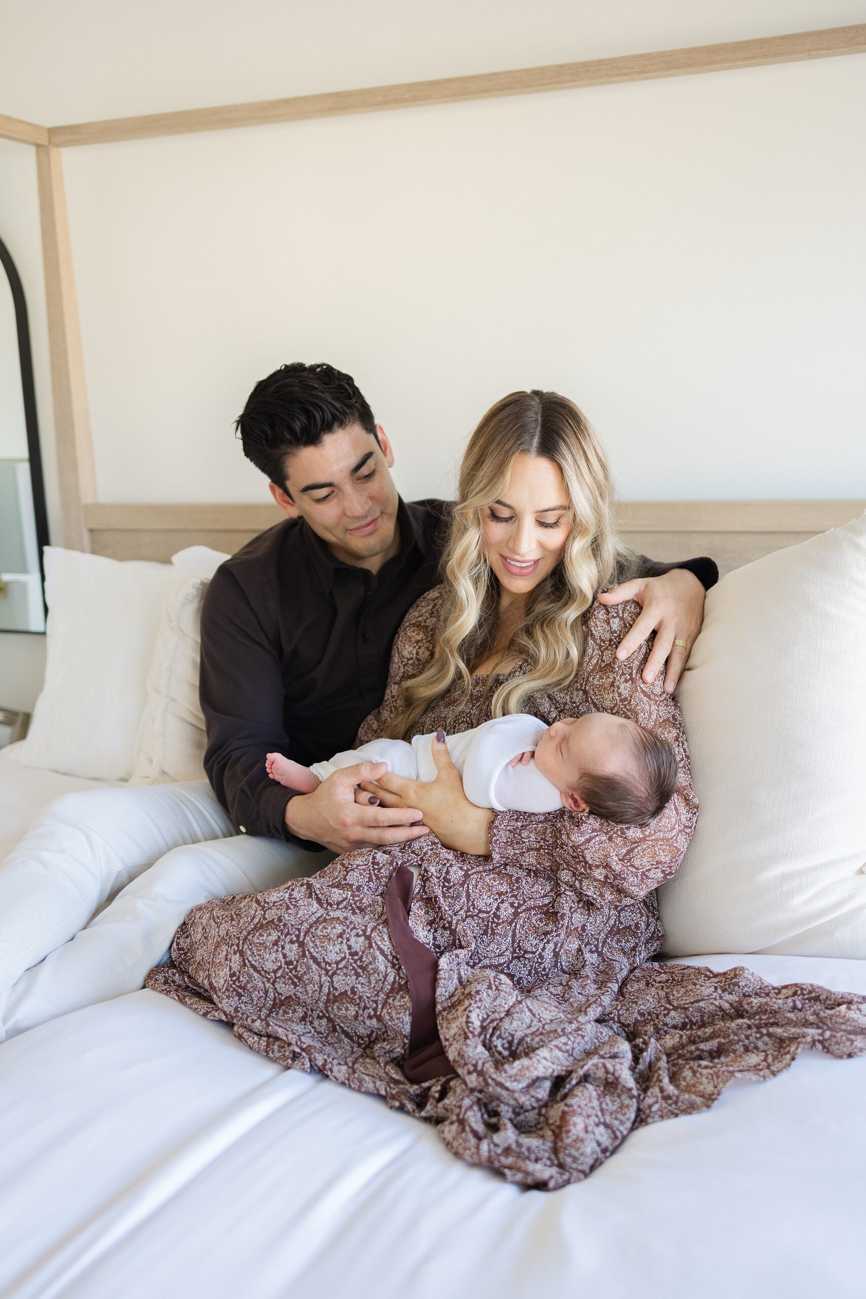 Happy new parents sit on a bed snuggling with their newborn baby in mom's arms after visiting yinRoot